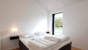 A modern bedroom with a large bed, a nightstand with a lamp, and a window with a view of greenery. - Holiday Home in Slagelse (Slagelse)