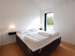 A modern bedroom with a large bed, a nightstand with a lamp, and a window with a view of greenery.