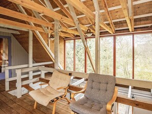Terraza de madera con dos sillas y una mesa, techo inclinado con vigas a la vista y ventana con vistas a árboles.