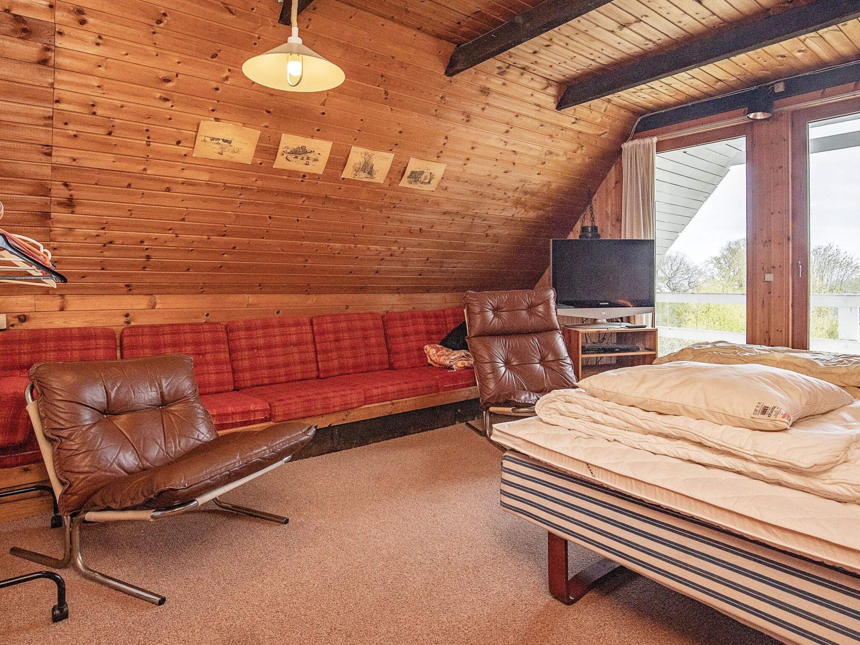 Ein gemütliches Zimmer mit einem roten Sofa, einem Ledersessel, einem Bett mit gestreifter Kopfteil, einem Fernseher und einer Holzzieldecke mit gerahmten Bildern.
