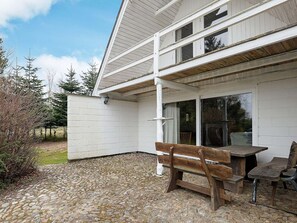 Une terrasse couverte avec un banc et une table en bois, des murs extérieurs blancs et un sol carrelé en pierre.