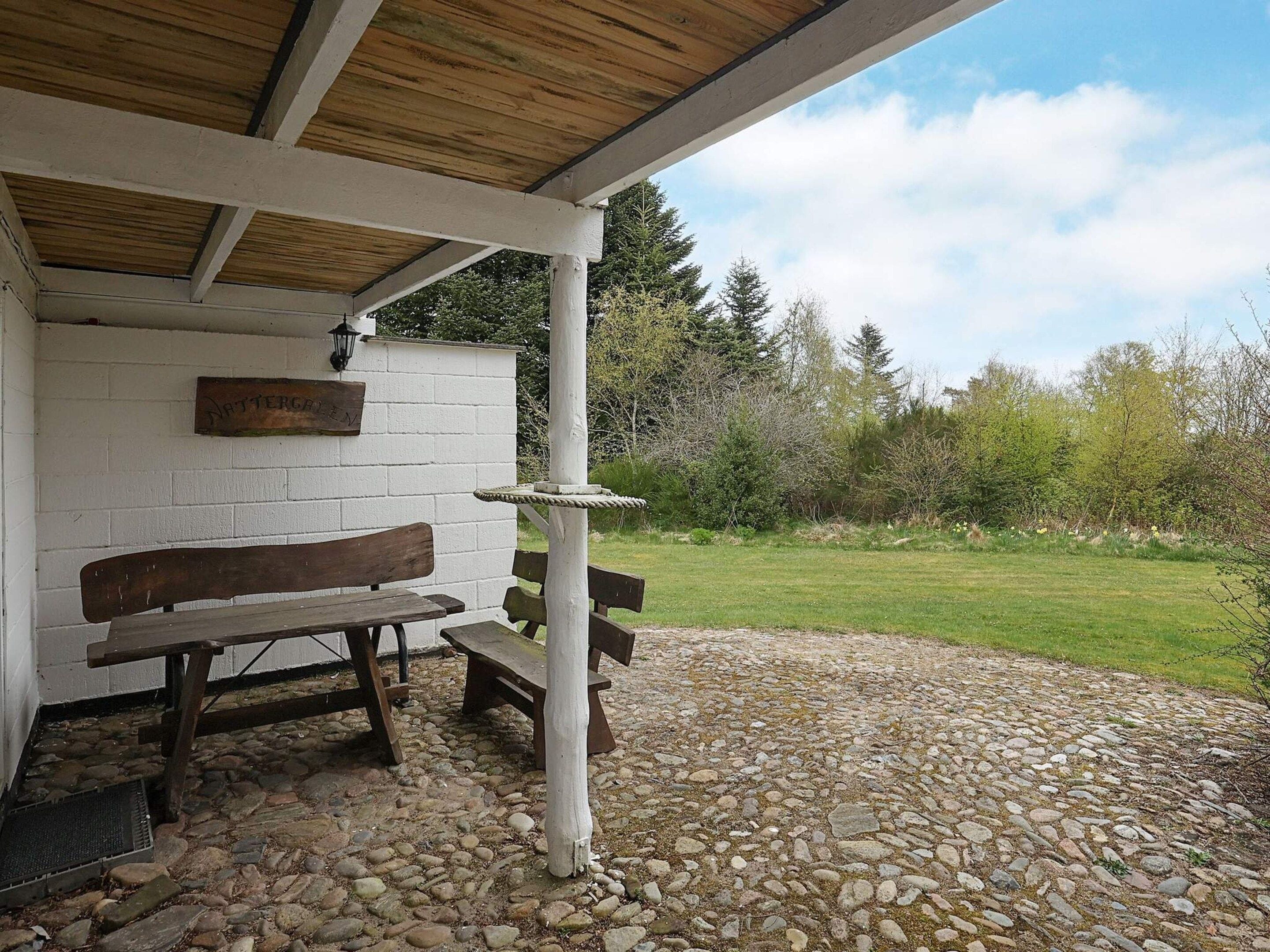 A covered outdoor seating area with a wooden bench and a stone-paved ground.