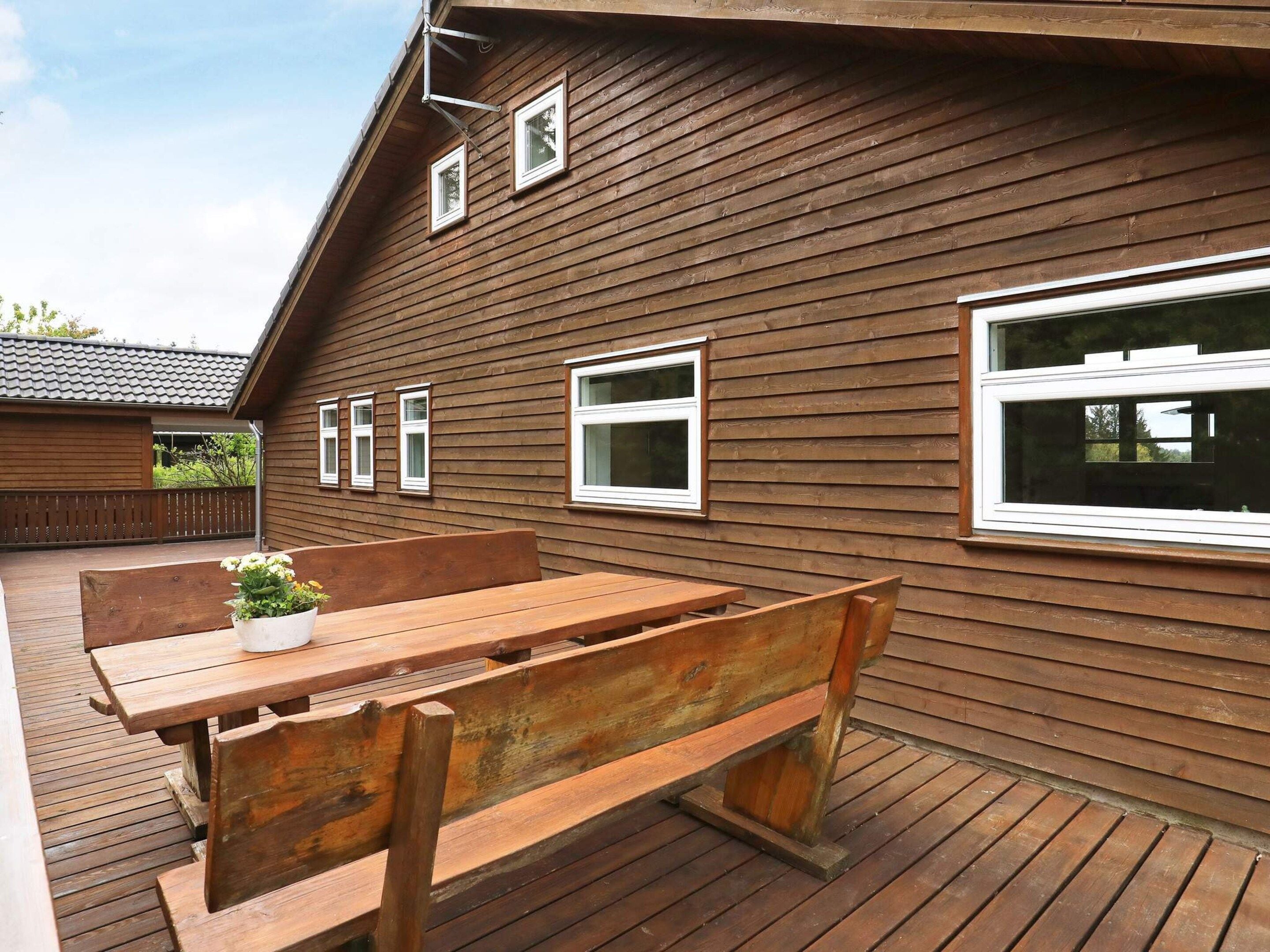 Una terraza de madera con un banco y una mesa, una casa de madera con varias ventanas, y un área cercada.