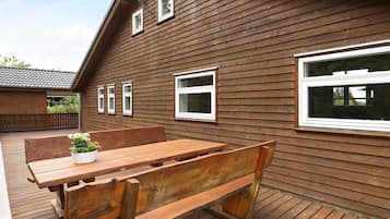 En træterrasse med træbænk og bord, et træhus med mange vinduer, og et indhegnet område.