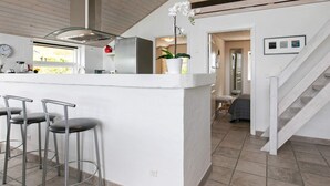 A modern kitchen with a central island, bar stools, a staircase, and a view of the adjacent living area. - Holiday Home in Skals (Skals)