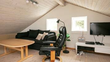 Ein modernes Schlafzimmer im Loftsstil mit schrägem Dach, einem schwarzen Ledersessel, einem Holztisch, einer schwarzen Couch, einem Fernseher und einem Schreibtisch mit Computer und Spielkonsole.