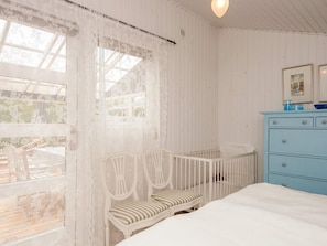 Quarto com cama, cômoda, janela com cortinas de renda e vista para um deque externo.