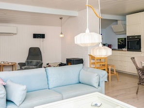 Uma sala de estar com sofá, poltrona, mesa, cozinha com armários e forno, e um lustre.