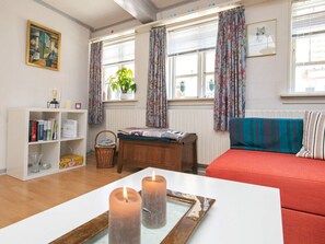 A cozy living room with a red sofa, a wooden table with candles, a bookshelf, and a window with curtains.