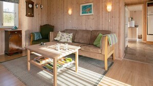 A cozy living room with a wooden floor, a sofa, a coffee table with magazines, a clock, and a painting on the wall. - Cozy Seaside Cottage Escape-by Traum (Fjerritslev)