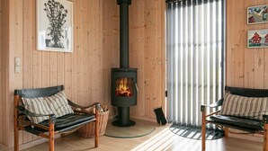 A cozy room with wooden walls, a central wood-burning stove, two leather chairs, a woven basket, and a striped rug. - Cozy Seaside Cottage Escape-by Traum (Fjerritslev)