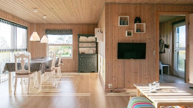 A wooden interior with a dining area, a TV mounted on the wall, and a cozy seating area with a colorful cushion. - Cozy Seaside Cottage Escape-by Traum (Fjerritslev)