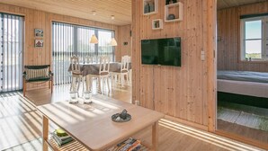 A spacious room with wooden walls, a dining area with a table and chairs, a bed, and a television mounted on the wall. - Cozy Seaside Cottage Escape-by Traum (Fjerritslev)