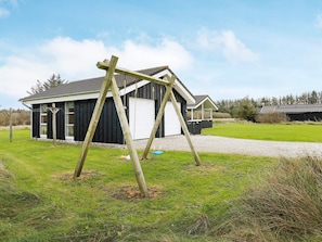 A black building with a white garage door, a swing set, and a gravel driveway. - Holiday Home in LÃ¸kken (Lokken)