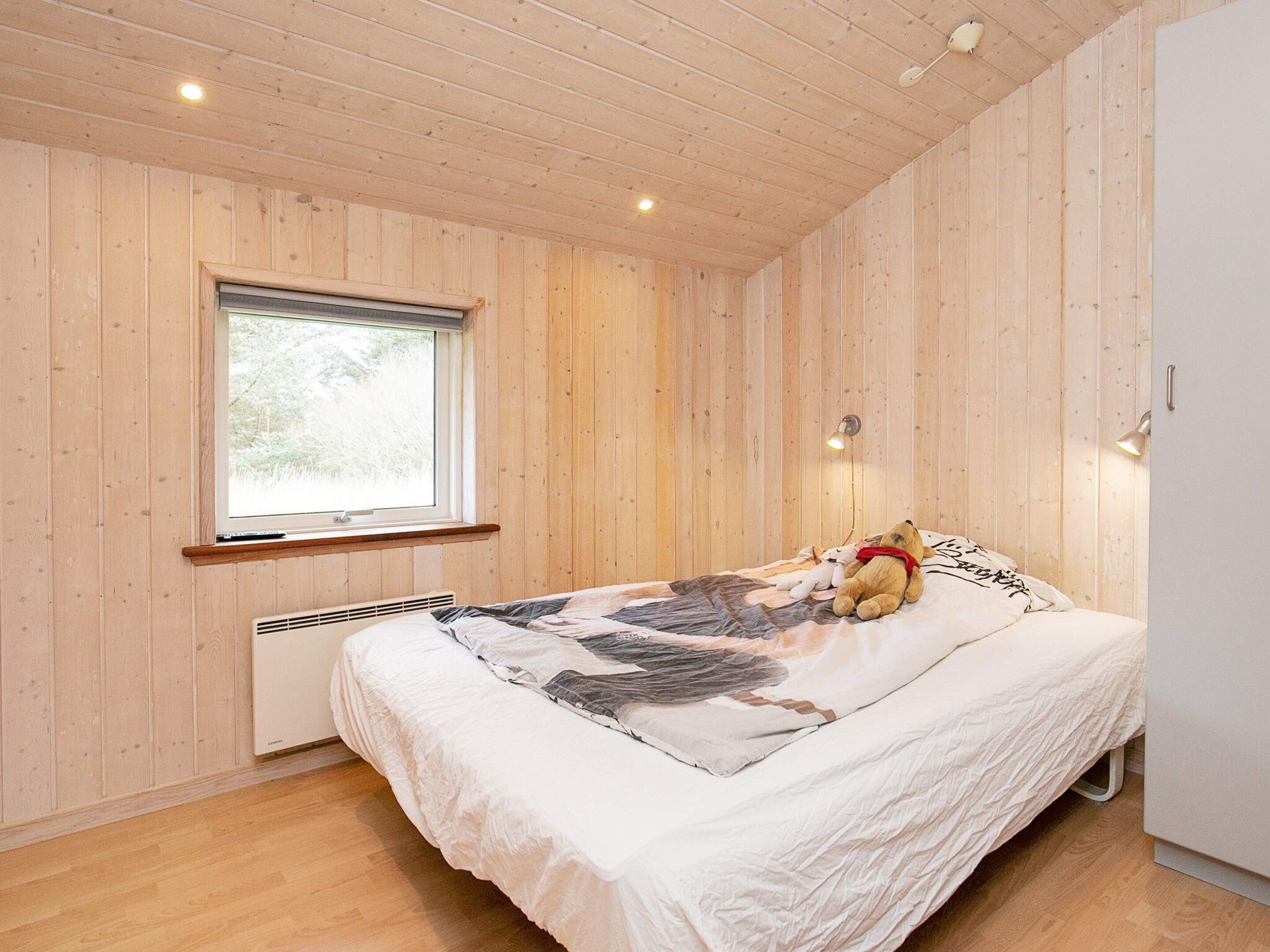A bedroom with wooden walls, a window, a bed with a patterned bedspread, a teddy bear, and a bedside lamp.