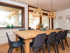 A dining area with a wooden table, six chairs, two hanging wicker lamps, and a window with potted plants.
