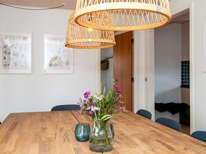 A wooden dining table with a vase of flowers, a hanging rattan light fixture, and framed botanical illustrations on the wall.