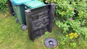 Ein grüner Müllcontainer mit blauem Deckel, ein Holzzaun und ein Metallgitter auf dem Boden.