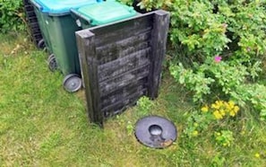 En grön soptunna med ett blått lock, ett trästaket och ett metallgaller på marken.