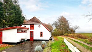 A white building with a red door and windows, surrounded by a white picket fence, a paved area, and a grassy field. - Cozy Coastal Cottage at Jegindo-by Traum (Thyholm)