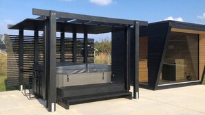 Un área moderna de hidromasaje al aire libre con una estructura de pérgola, una terraza de madera y un edificio con techo inclinado.