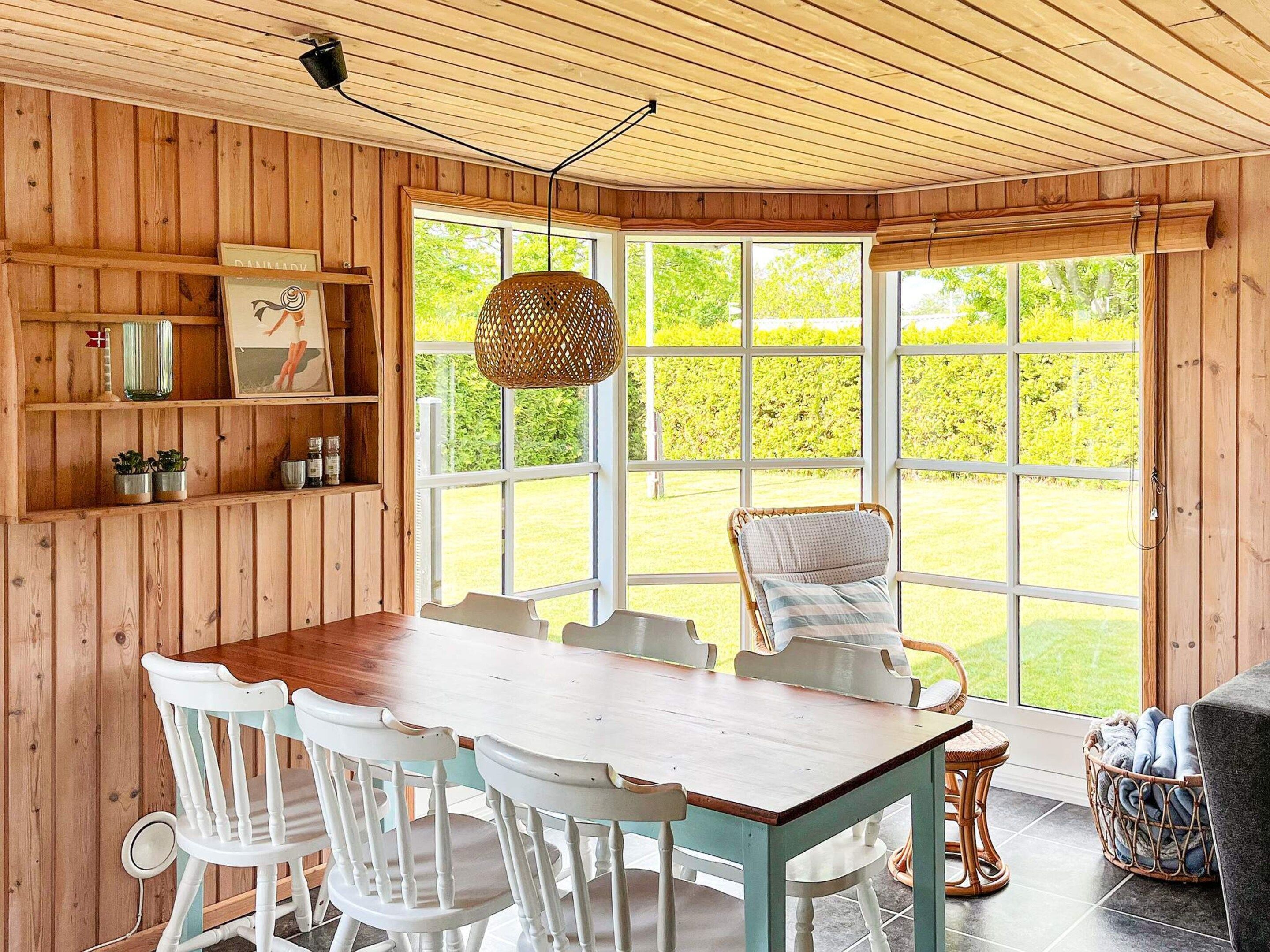 Una habitación con paneles de madera, una mesa de comedor con sillas, una lámpara colgante de mimbre y un ventanal con vistas a la vegetación.