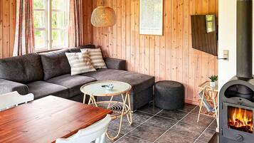 Une pièce chaleureuse avec un canapé, une petite table ronde, une chaise, une table en bois, un luminaire tressé, un tableau encadré et un poêle à bois.