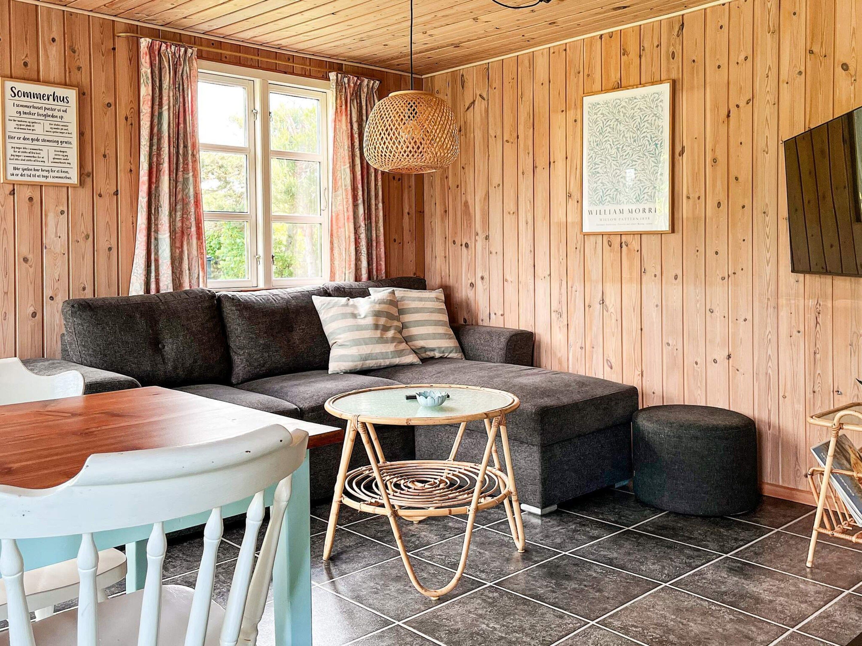 A cozy room with a sofa, a round table, and a chair. There's a window with curtains, a hanging light fixture, and a framed picture on the wall.