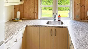 A kitchen with wooden cabinets, a granite countertop, a stainless steel sink, and a window with a view of a green lawn. - Holiday Home in Hadsund (Hadsund)