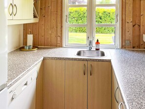 A kitchen with wooden cabinets, a granite countertop, a stainless steel sink, and a window with a view of a green lawn. - Holiday Home in Hadsund (Hadsund)