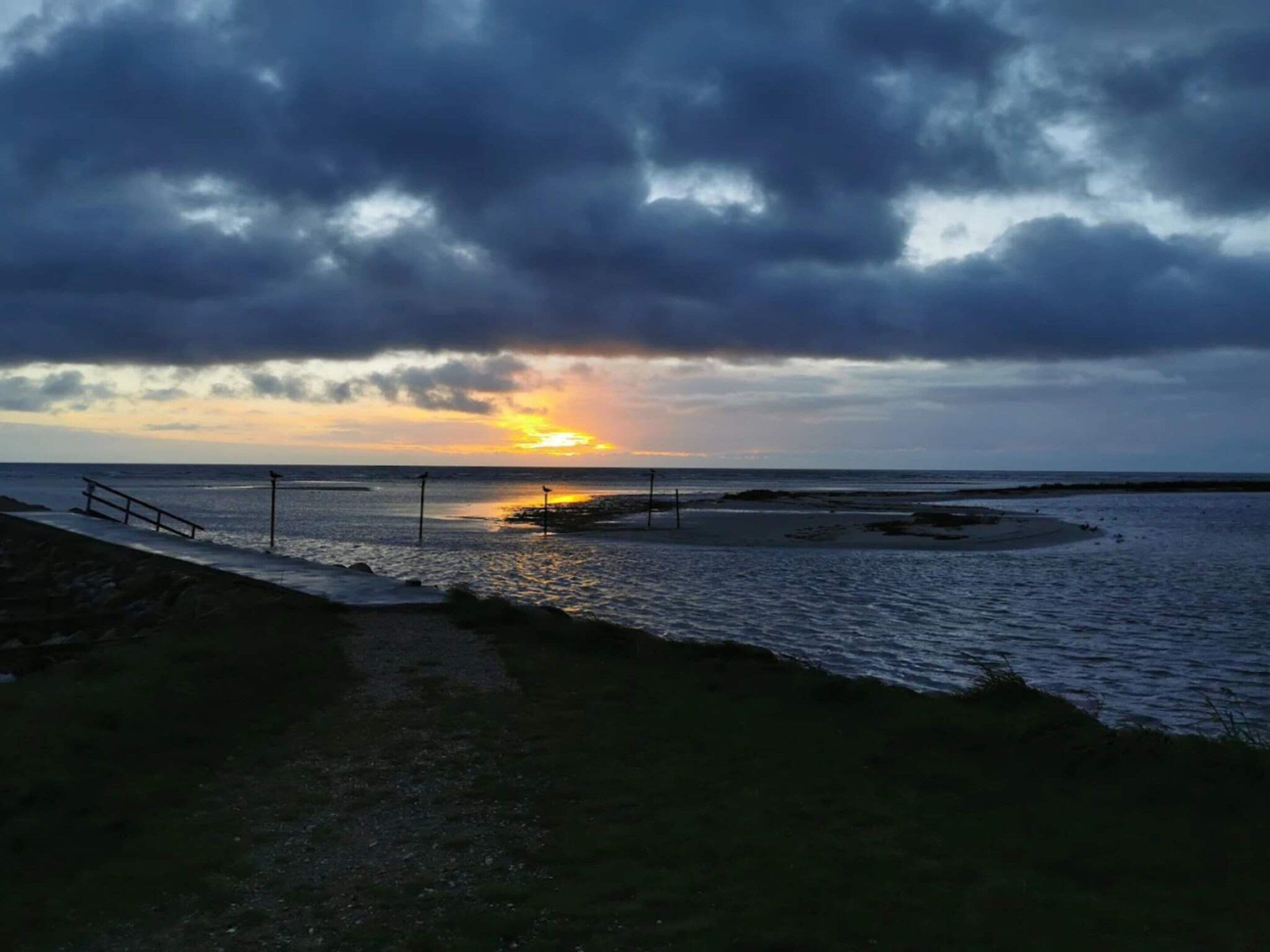 Solnedgang ved kysten med strand og træpromenade.