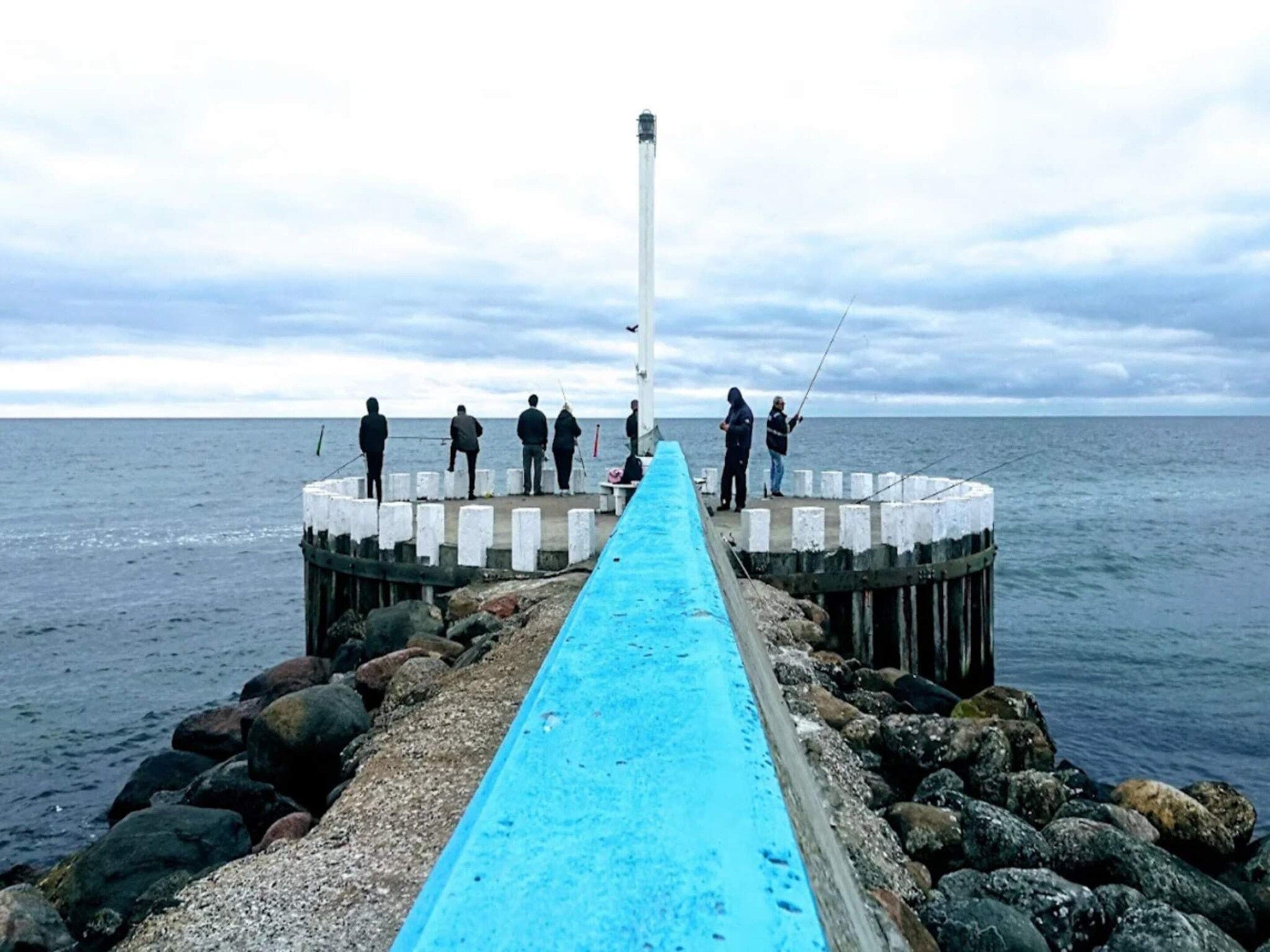 A pier extending into the water with a blue walkway, surrounded by rocks and a lighthouse in the background.