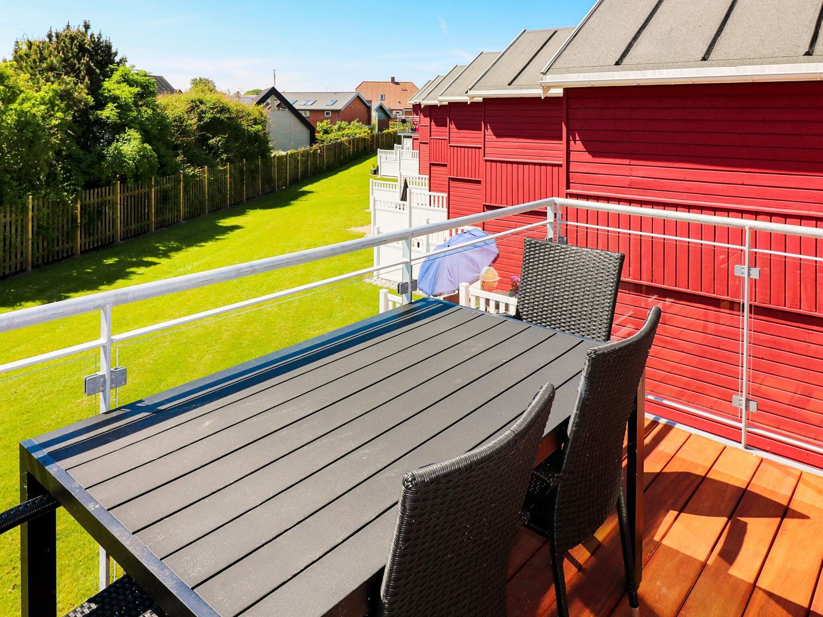 A balcony with a table and chairs, a red building in the background, and a green lawn.