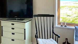 Quarto com televisão sobre um móvel, uma cadeira com vista para a praia e uma janela com vista para a praia.