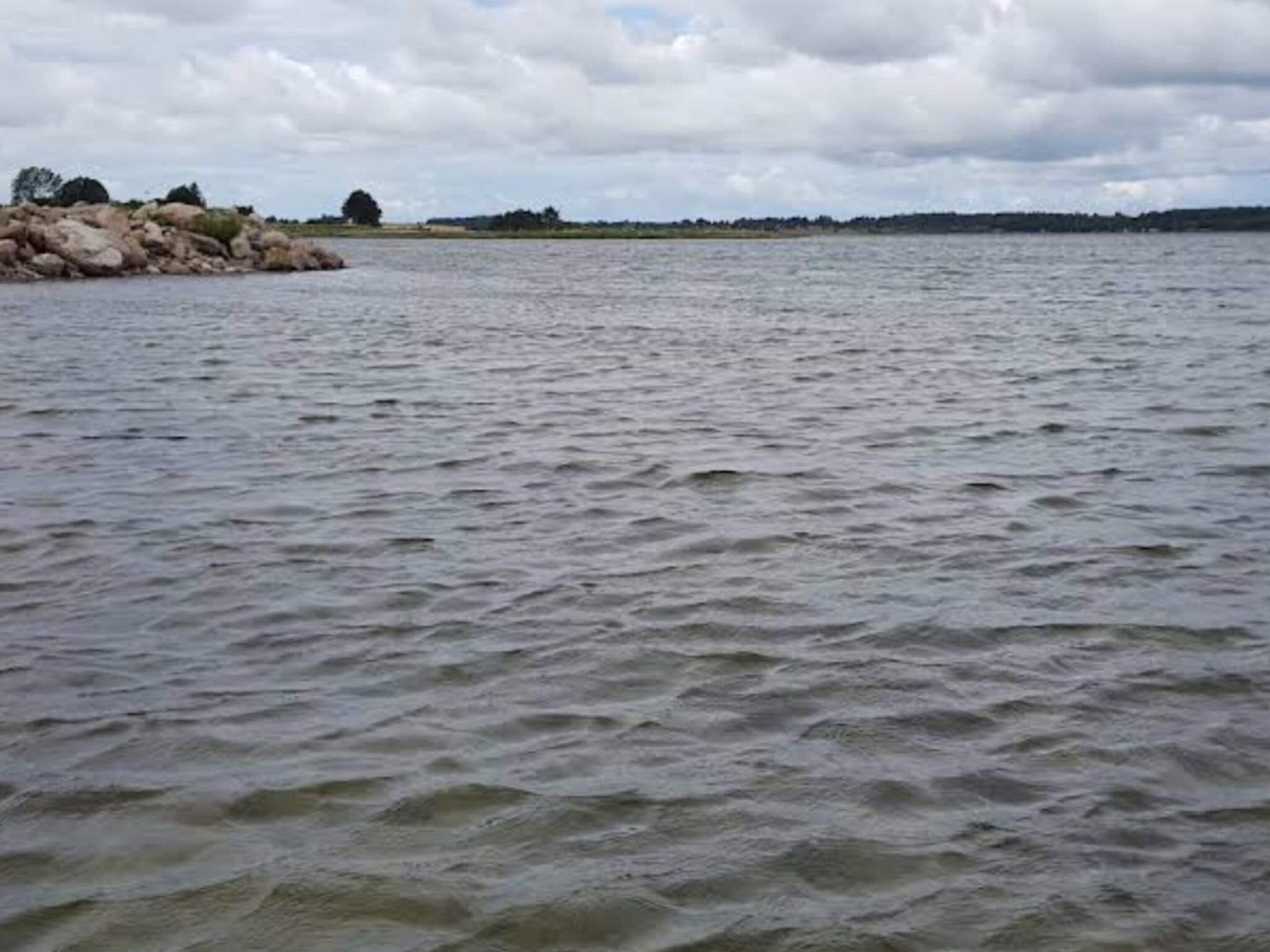 A calm body of water with a rocky shoreline and distant trees under a cloudy sky.