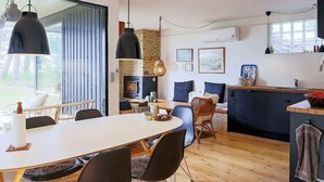 A modern kitchen with a central island, a dining area with a white table and black chairs, and a cozy seating area with a wicker chair and cushions. - 5 Person Holiday Home in Jaegerspris (Jægerspris)