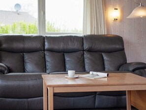 A room with a black leather sofa, a wooden coffee table with a cup and a magazine, and a window with curtains.