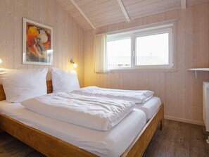 Een kamer met houten wanden en een groot bed met witte beddengoed. Er is een raam met gordijnen, een schilderij aan de muur en een radiator aan de rechterkant.