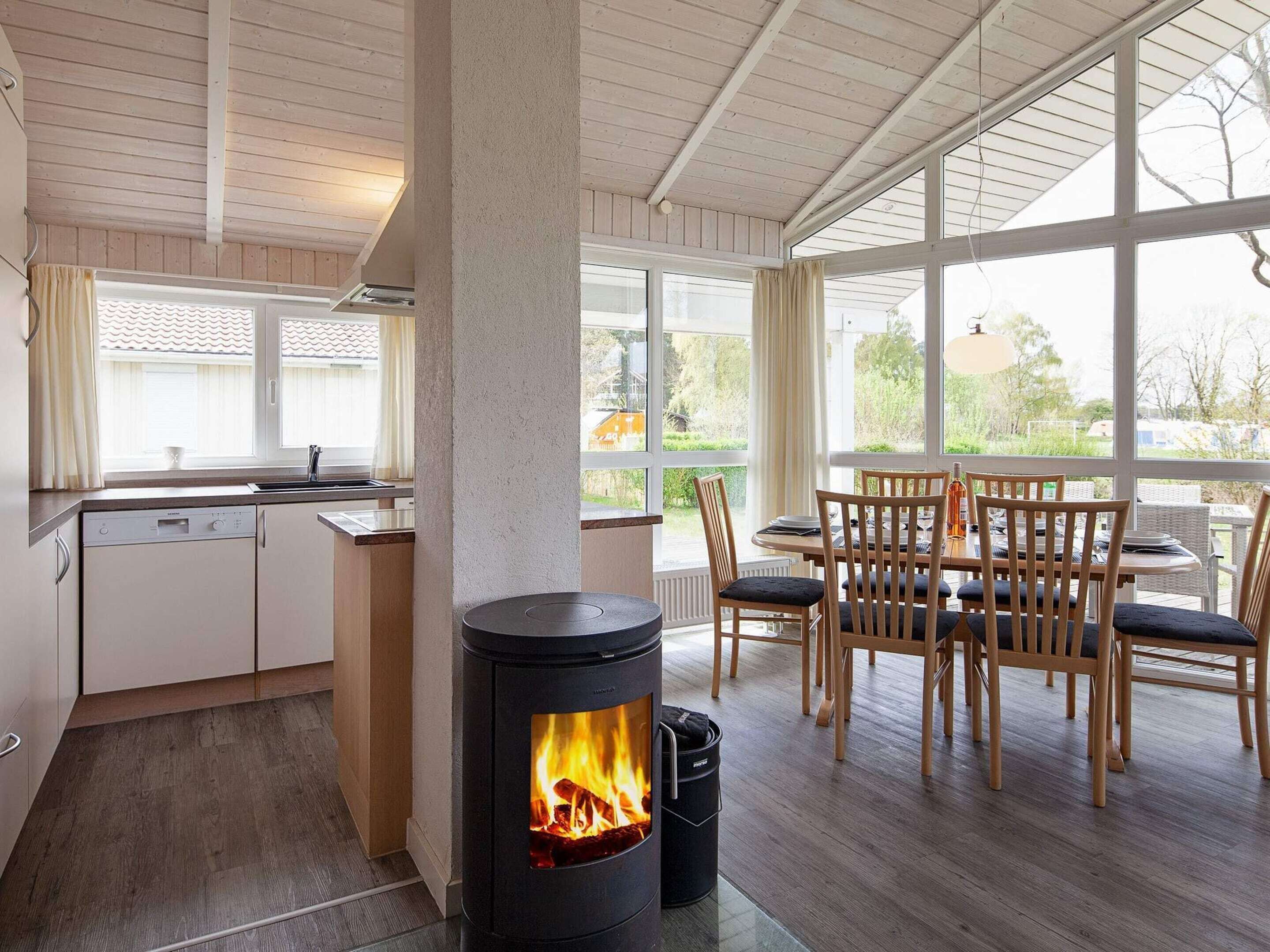 A modern kitchen with a wood-burning stove, a dining area with wooden chairs, and large windows offering a view of the outdoors.