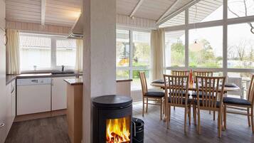 A modern kitchen with a wood-burning stove, a dining area with wooden chairs, and large windows offering a view of the outdoors.