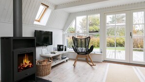 A cozy room with a fireplace, a television, a comfortable chair, and large windows offering a view of the outdoors.