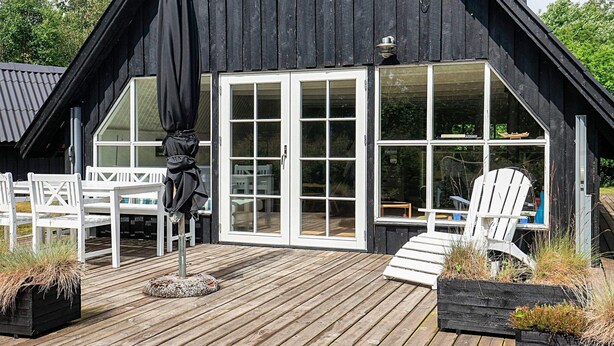 A wooden deck with white chairs and a table, a black umbrella, and a large window with a view of the interior.