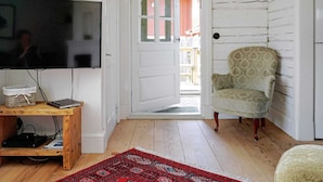 Ein Zimmer mit Fernseher auf einem Holztisch, einem gemusterten Sessel, einem roten Teppich und einer weißen Tür, die nach draußen führt.