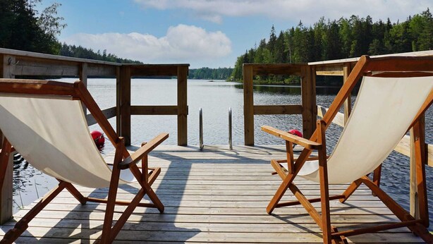 Eine Holzterrasse mit zwei Korbstühlen, die auf einen ruhigen See blicken, umgeben von Bäumen und einem klaren Himmel.