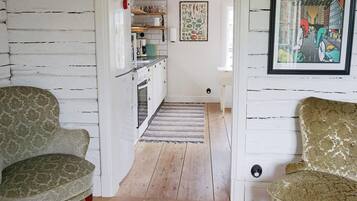 A hallway with two patterned armchairs, a kitchen area with wooden beams, and a framed poster on the wall.