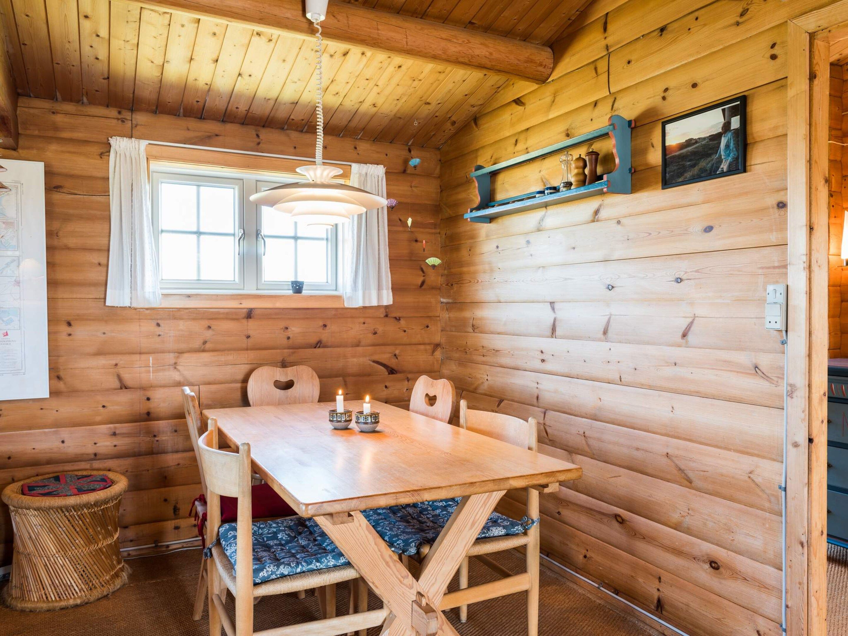 A wooden cabin interior with a dining table, chairs, a hanging light fixture, a window, and a shelf with decorative items.