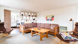 Uma sala de estar aconchegante com um sofá de madeira, uma mesa de centro de madeira e uma poltrona de couro marrom. Uma cozinha com armários brancos e prateleiras de madeira é visível ao fundo.