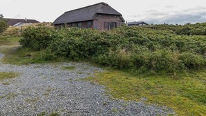 Uma casa rústica com telhado de palha, rodeada por vegetação densa e um caminho de cascalho que a leva.