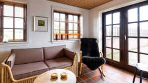 Um quarto aconchegante com um sofá xadrez, uma poltrona de vime, uma mesa de madeira redonda com velas e janelas amplas com persianas.