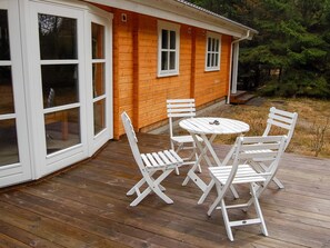 Una terraza de madera con una mesa y sillas blancas, un porche cubierto y una casa con revestimiento naranja y detalles blancos.